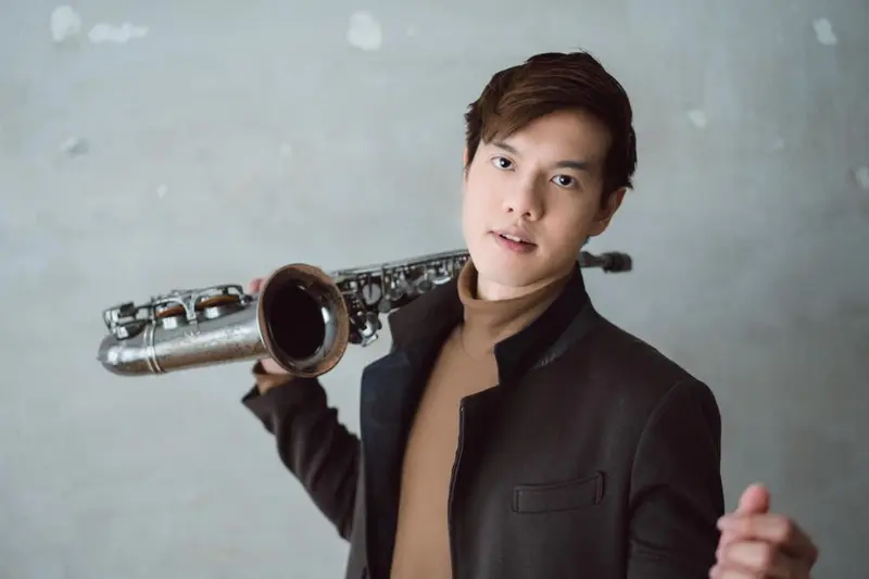 Saxophonist holding his saxophone over his shoulder against a grey wall