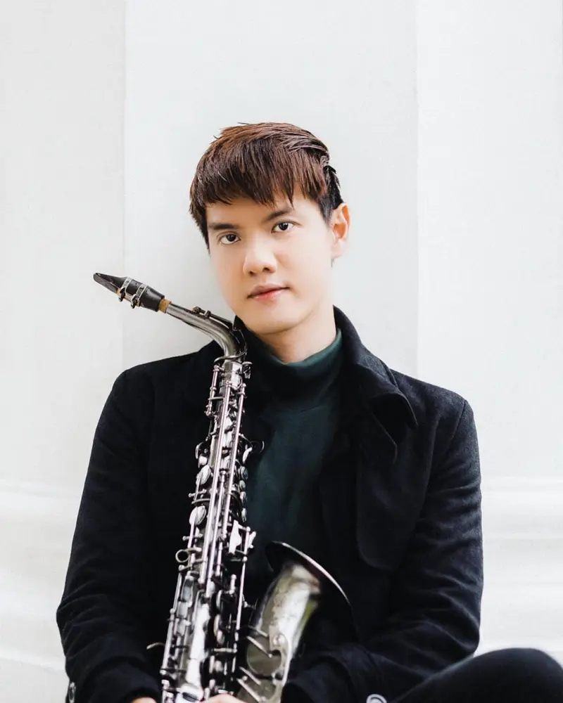Saxophonist in a black coat sitting with his saxophone against a white wall