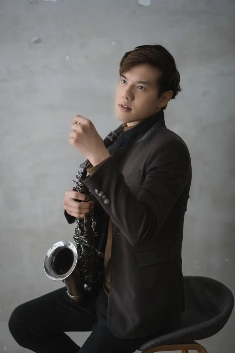 Saxophonist in a brown jacket seated on a stool holding an alto saxophone