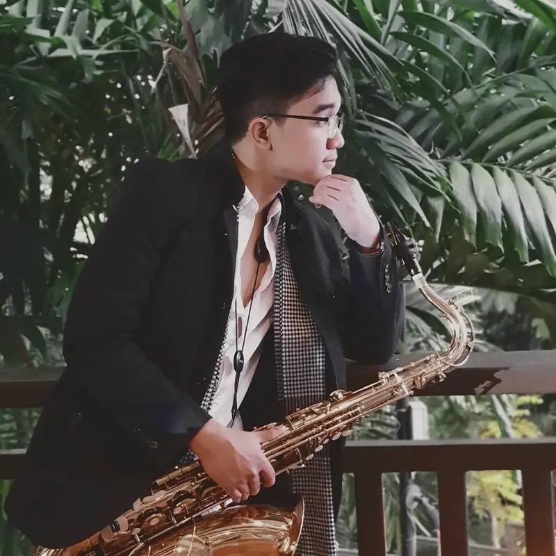 Saxophonist in a dark blazer holding a gold tenor saxophone on a terrace with palm leaves