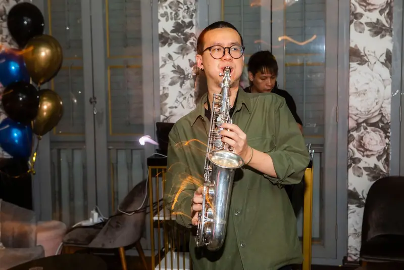 Saxophonist playing a silver alto sax with the DJ behind him at an evening party