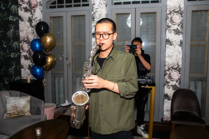 Saxophonist playing a silver saxophone at a party while a guest films on a phone